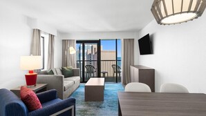 Une chambre d’hôtel équipée d’un canapé, d’un fauteuil, d’une table basse et d’une table à manger. Un téléviseur est fixé au mur et il y a un balcon avec des chaises.