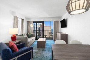 A hotel room with a sofa, armchair, coffee table, and a dining table. There is a TV mounted on the wall and a balcony with chairs.
