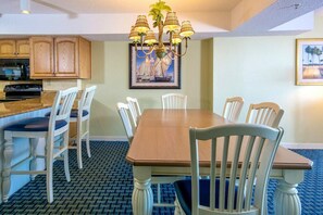 Uma área de refeições com mesa de madeira e cadeiras brancas, um lustre e uma cozinha ao fundo.