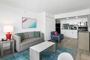 Uma sala moderna com um sofá cinzento, uma mesa de centro branca e um tapete azul. Próximo, encontra-se uma cozinha compacta com armários brancos e um micro-ondas.