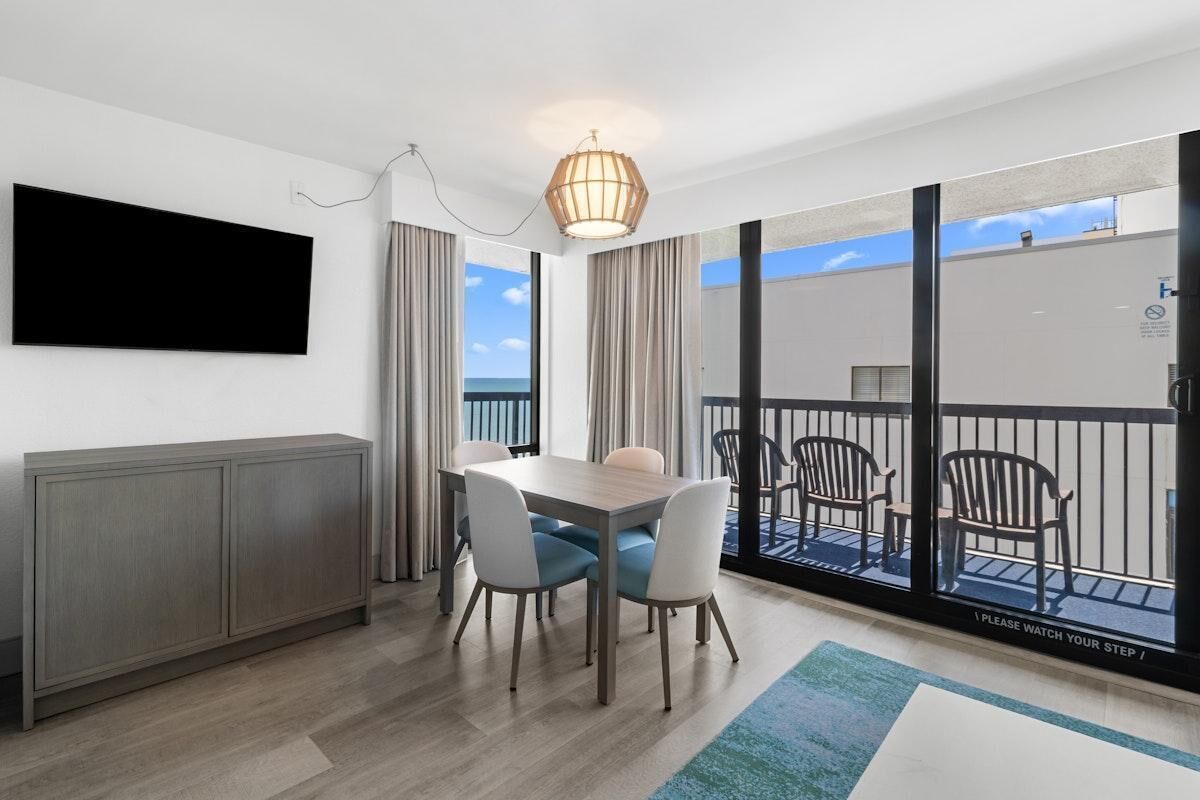 Una habitación moderna con una mesa de comedor, sillas, un televisor montado en la pared y un balcón con mobiliario exterior.