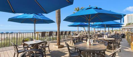 Une terrasse extérieure avec des tables et des chaises, des parasols bleus et une vue sur l’océan.