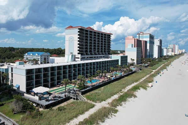 A hotel with a modern design, featuring a large pool area, a beachside location, and multiple buildings in the background.