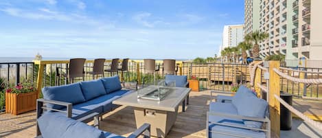 A rooftop terrace with outdoor seating, a fire pit, and a view of the ocean and buildings.
