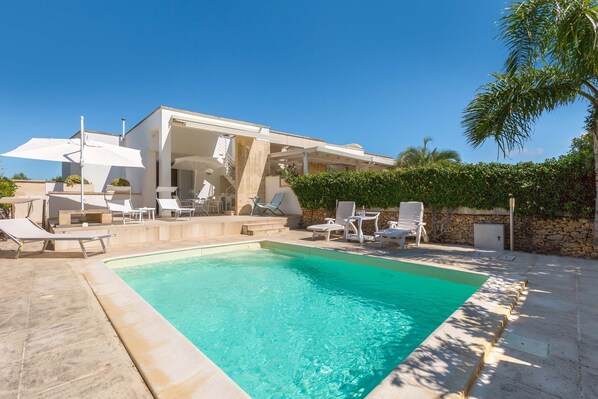 A modern pool area with a rectangular pool, white lounge chairs, and a covered seating area with umbrellas.
