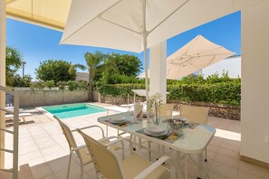 A patio with a dining table, chairs, and umbrellas, overlooking a swimming pool and greenery.