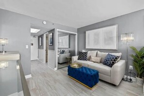 A modern living room with a white sofa, blue ottoman, and a large abstract painting on the wall.