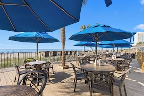 An outdoor patio with tables and chairs, blue umbrellas, and a view of the ocean.