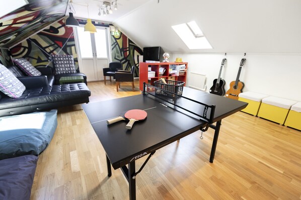 Una habitación con mesa de ping pong, sofá, estantería y guitarra.