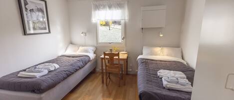 Une petite pièce bien éclairée, avec deux lits simples, une table en bois et une fenêtre donnant sur l’extérieur.