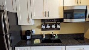 A kitchen with white cabinets, a stainless steel refrigerator, a microwave, a coffee station, and a sink with soap dispensers. - Ocean Therapy (Carolina Beach)