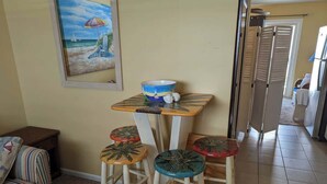 A dining area with a table and four stools, a painting on the wall, and a kitchen in the background. - Ocean Therapy (Carolina Beach)