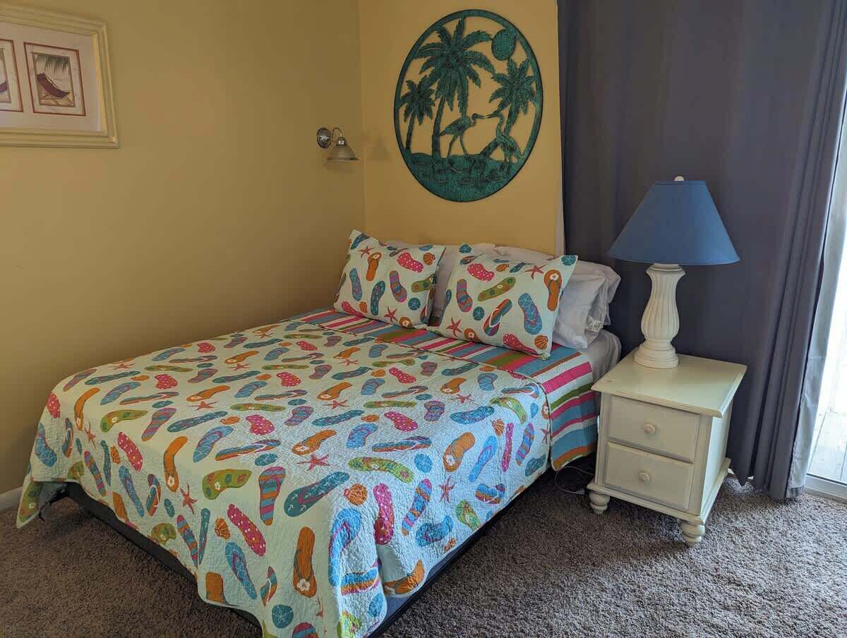 Une chambre à coucher comprenant un lit, une table de chevet, une lampe, un élément de décoration murale et un sol recouvert d’une moquette.