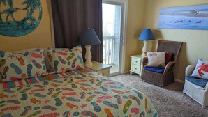 A bedroom with a bed, two chairs, a nightstand, a lamp, and a painting on the wall. - Ocean Therapy (Carolina Beach)
