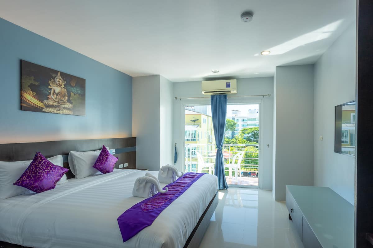 A modern hotel room with a large bed, a painting on the wall, a balcony with a view, and a ceiling fan.