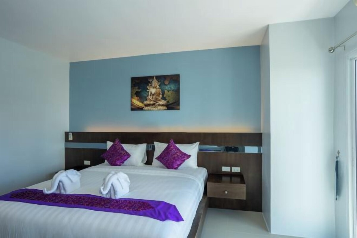 A neatly made bed with white linens and purple accents, a painting of a seated figure above it, and a wooden headboard.