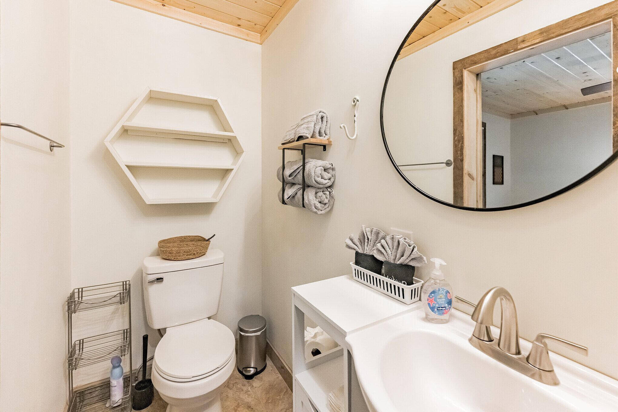A bathroom with a toilet, sink, mirror, towel rack, and storage shelves.