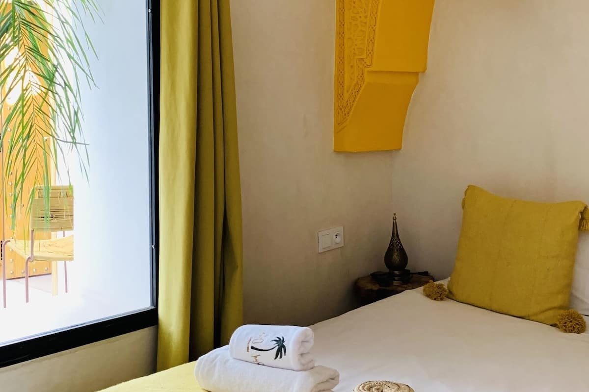 A hotel room with a bed, yellow curtains, a woven ceiling light, and a view of palm trees through the window.