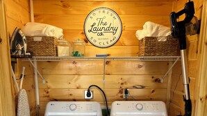Una sala de bugaderia amb dues rentadores LG blanques, una cistella de vimetes, un pot i un cartell que diu "The Laundry Co. Self-Serve Clean". 