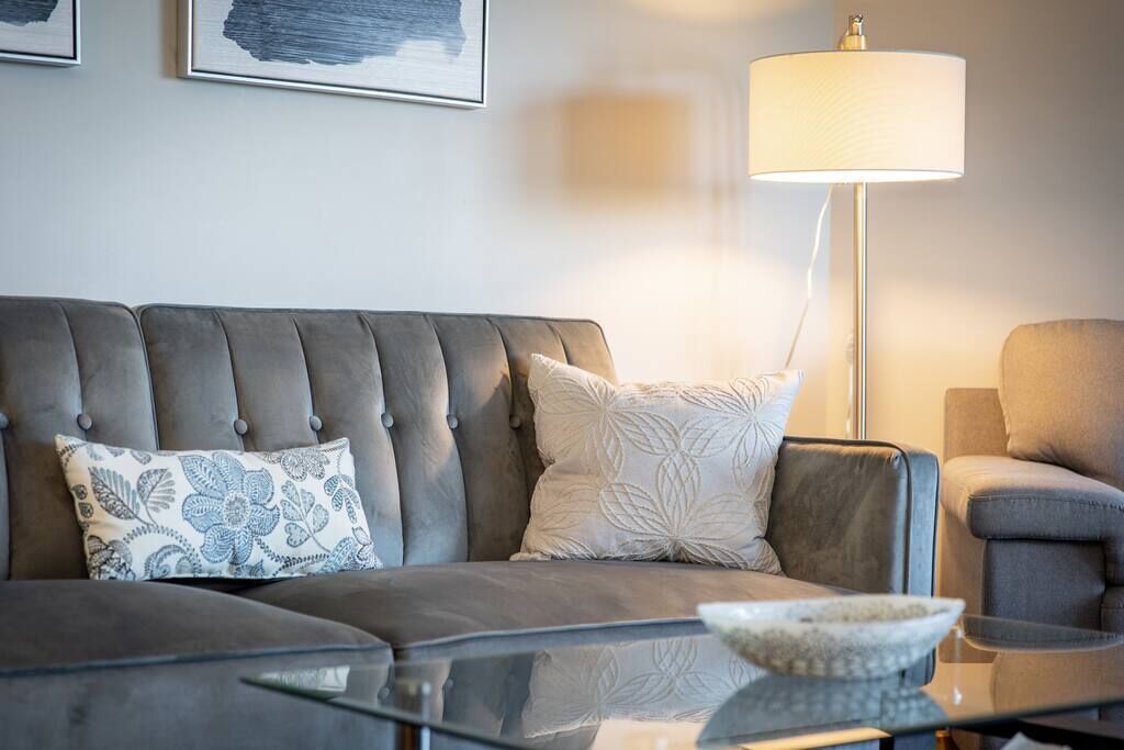 A modern living room with a grey sofa, two patterned cushions, a glass coffee table, and a floor lamp.