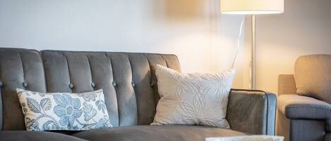 A modern living room with a grey sofa, two patterned cushions, a glass coffee table, and a floor lamp.