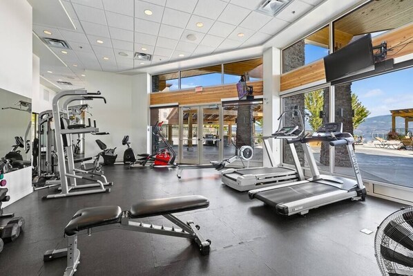 Un gimnasio con diversos equipos de ejercicio, ventanas amplias y vistas al exterior.