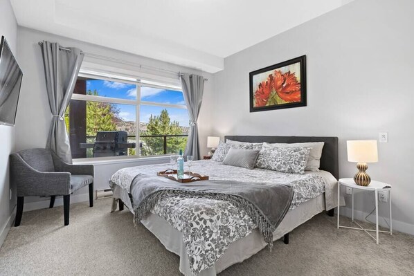 A modern bedroom with a large bed, a chair, a bedside table, and a window with curtains. - West Kelowna Family-friendly condo/AC/pool/Hot tub/winery/lake view (Central Okanagan)