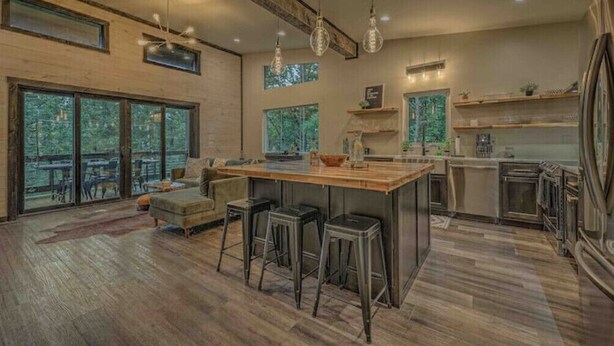 A spacious kitchen with a large island, bar stools, and a view of the outdoors through large windows. - Brand New Relaxing Mountain Retreat 
 (Talking Rock)
