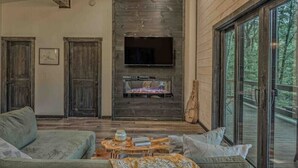 A modern living room with a fireplace, a television, and a large window overlooking a deck. - Brand New Relaxing Mountain Retreat 
 (Talking Rock)