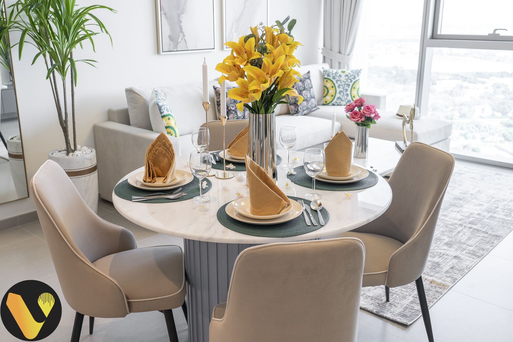 Un comedor con una mesa redonda dispuesta para cuatro personas, rodeada de cuatro sillas. Hay platos, servilletas y vasos sobre la mesa. Un jarrón con flores amarillas está en la mesa, y una planta en maceta se ve al fondo.