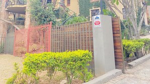 A two-story building with a yellow facade, a red fence, and a small garden with green shrubs. - Full Moon Guest House (Mbombela)