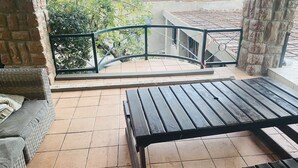 A balcony with a wicker sofa, a wooden table, and a view of a construction site with buildings and trees. - Full Moon Guest House (Mbombela)