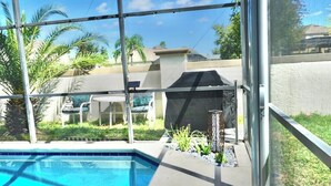 Una piscina con cerramiento de vidrio, un patio con mobiliario exterior y vistas a una zona residencial con palmeras y una casa.