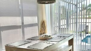 Un área de comedor moderna con una mesa de madera y bancos, ventanas grandes con persianas y vista a una piscina.