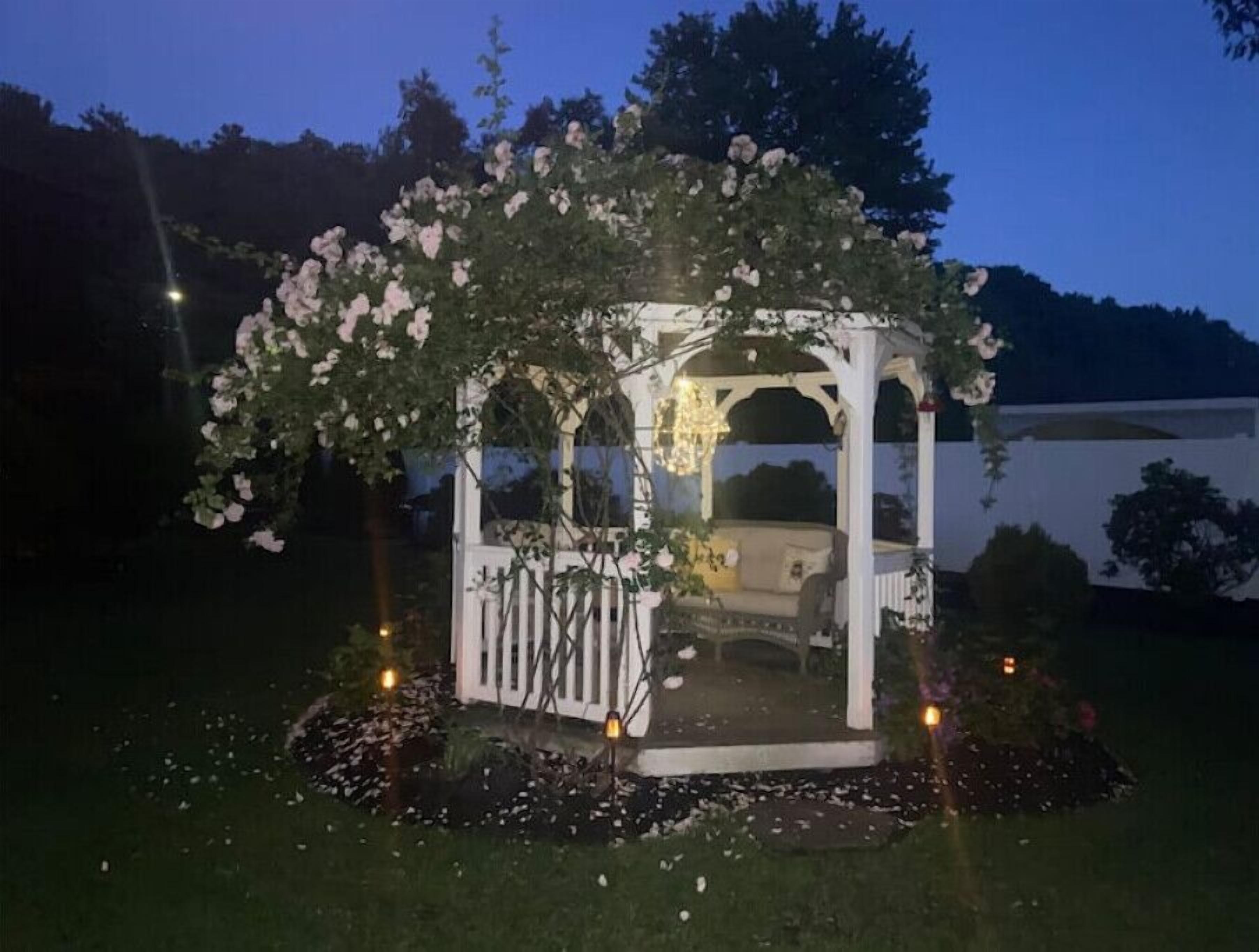 A gazebo with a white structure and a roof covered in white flowers, surrounded by a garden with scattered petals.