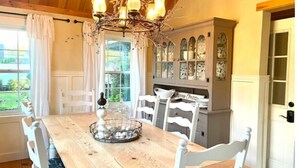 Une salle à manger avec une table en bois, des chaises blanches, un lustre et un buffet aux portes vitrées.