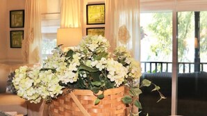Une pièce avec une grande fenêtre, une enseigne en bois au-dessus sur laquelle on peut lire « ANTIQUITÉS », et un panier de fleurs blanches posé sur une table.