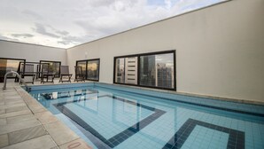 A swimming pool with tiled flooring, a seating area with chairs, and large windows offering a city view. - Estúdio Aconchegante, Metrô, Piscina, Sauna, AC (São Paulo)