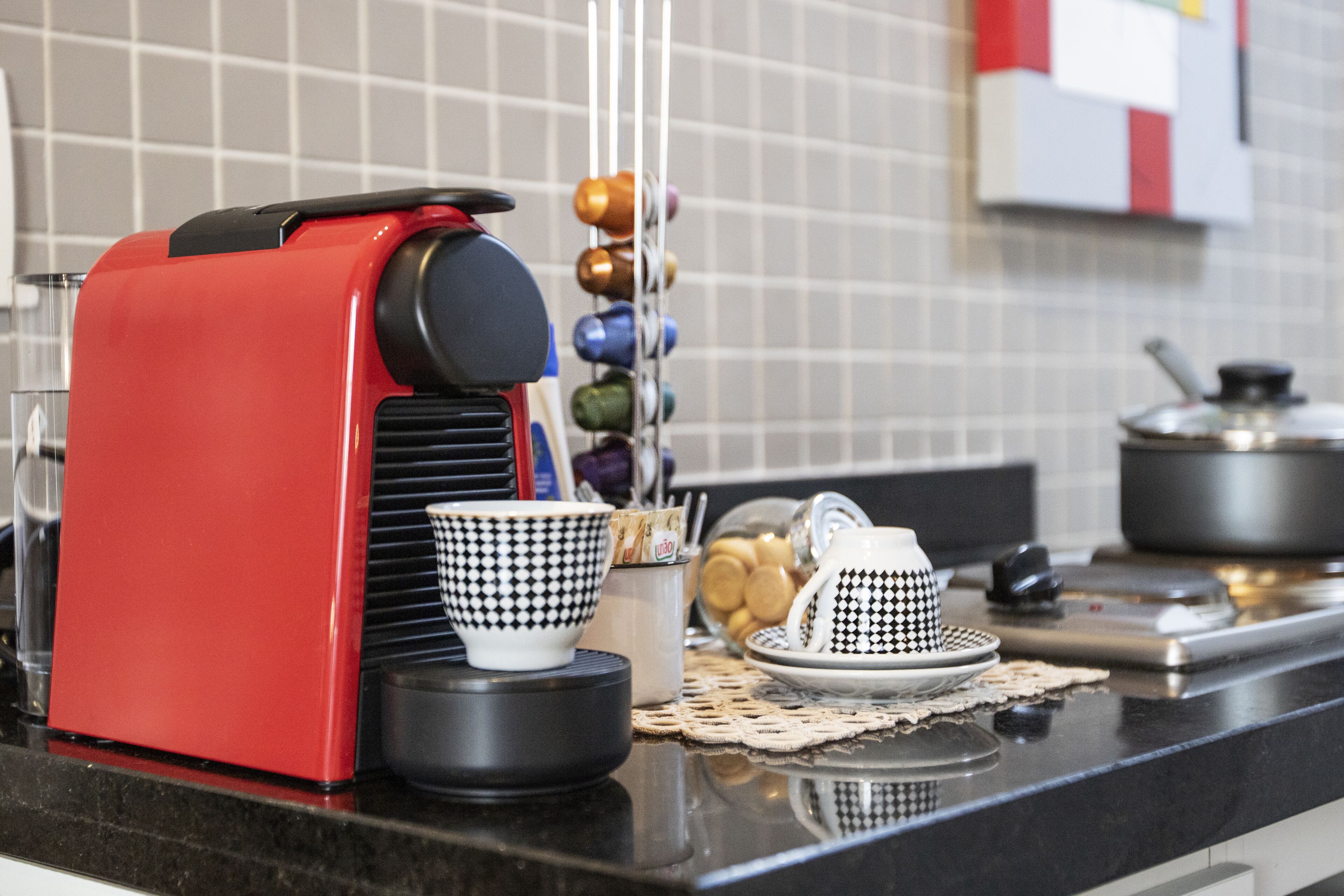 Un comptoir de cuisine avec une machine à expresso rouge, des tasses à carreaux noirs et blancs, et une variété de tasses colorées.