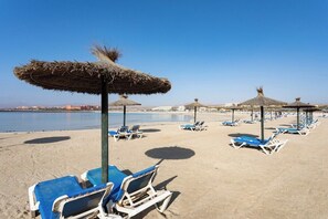 A beach with rows of blue lounge chairs and thatched umbrellas, a clear sky, and a calm sea in the background.