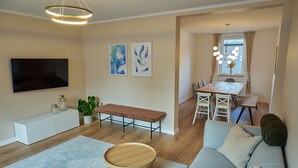 Ein modernes Wohnzimmer mit einer Couch, einer Bank, einem Esstisch mit Stühlen, einem an der Wand befestigten Fernseher und abstrakten Kunstwerken.