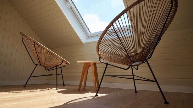 Ein Zimmer mit zwei Korbstühlen, einem kleinen Holztisch, einem Oberlicht und Holzböden.