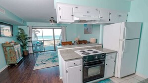 Une cuisine équipée d’une cuisinière, d’un four et d’un réfrigérateur, un coin repas avec des chaises, et une vue sur l’océan par une porte coulissante.