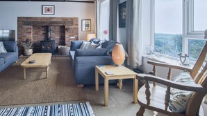 A cozy living room with a brick fireplace, a sofa, a wooden coffee table, and a view of the outdoors through a large window.