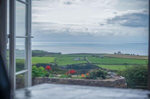 Vista desde una ventana sobre un paisaje costero con campos y una casa a lo lejos.