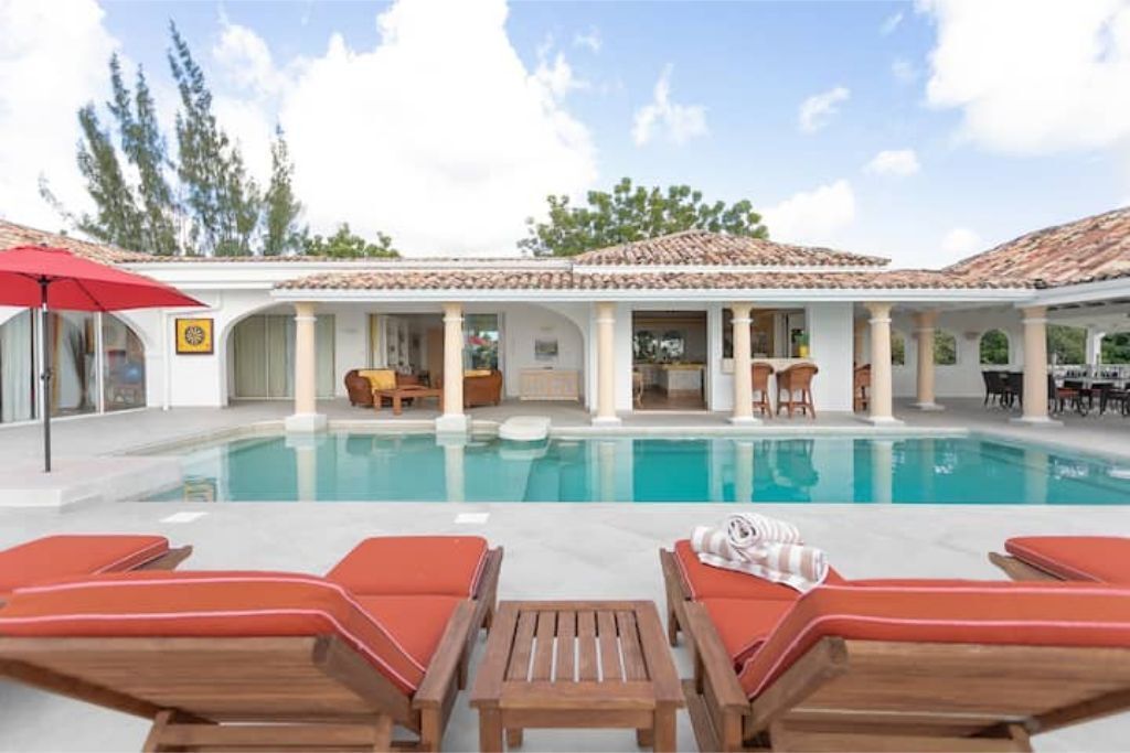 A poolside area with lounge chairs and a table, a covered patio with chairs, and a clear blue pool.