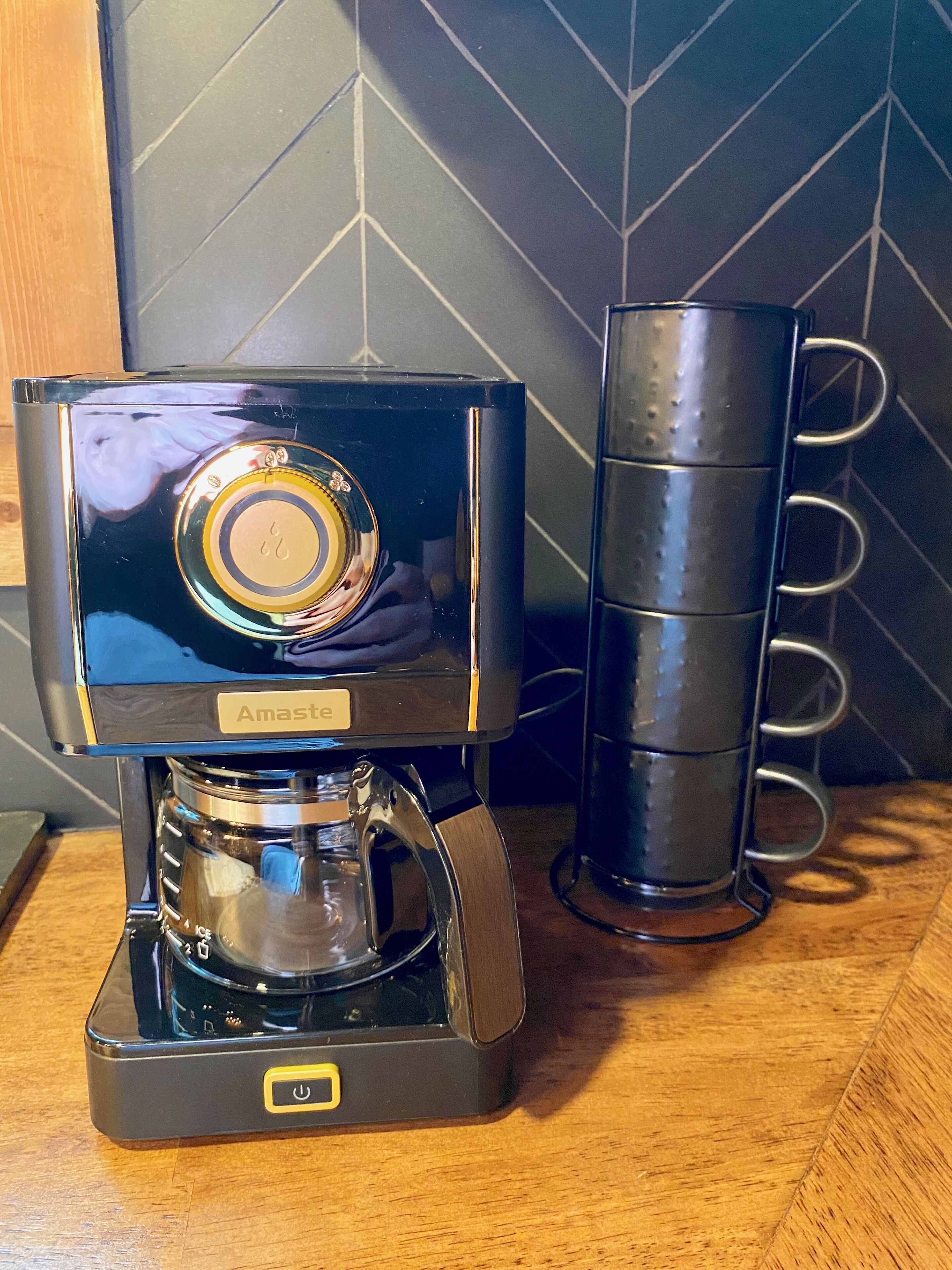 A coffee maker with a glass carafe and a stack of black mugs on a wooden surface.