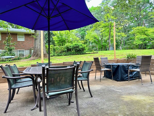 An outdoor patio with a table and chairs, a blue umbrella, and a view of a grassy area with trees.