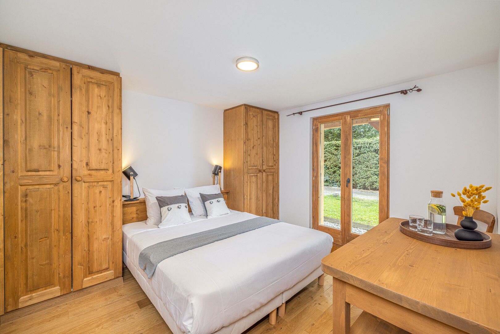 A bedroom with a bed, wooden wardrobe, bedside lamps, a table with a vase, and a view through a glass door.