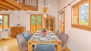 Comedor con mesa de madera dispuesta para seis personas, rodeada de sillas. Se observa un aparador de madera con puertas de cristal, un reloj en la pared y vistas al exterior con vegetación.